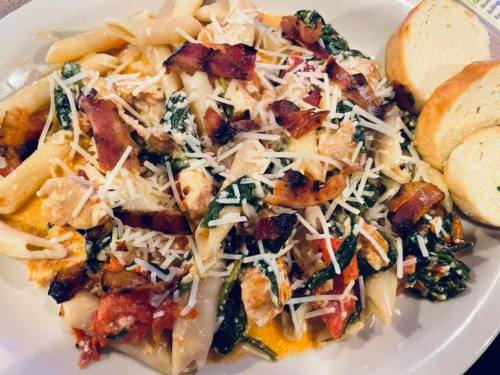 A plate of pasta with chicken, spinach, tomatoes, bacon, and cheese, served with two slices of bread.