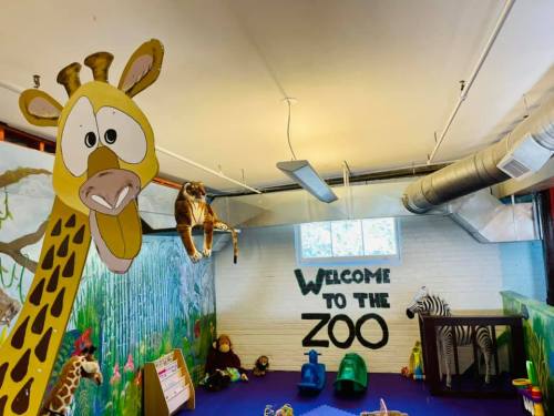 Colorful indoor play area with a giraffe mural, toy animals, and a "Welcome to the Zoo" sign on the wall.
