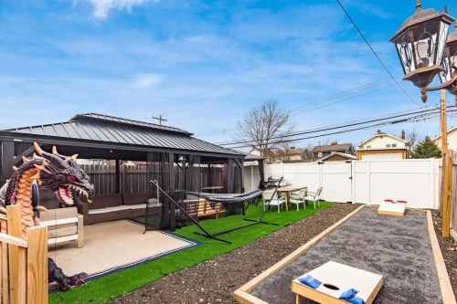 Backyard with a gazebo, hammock, cornhole game, and a dragon statue, surrounded by a fence and blue sky.