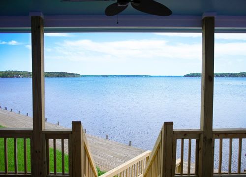 View of a calm lake from a porch, with wooden steps leading down to a dock and lush greenery in the background.