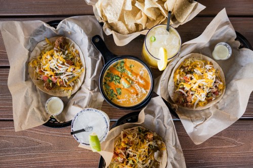A spread of tacos with toppings, a bowl of orange sauce, chips, and drinks on a wooden table.