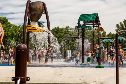 Children playing in a splash pad with water features and playground structures on a sunny day.