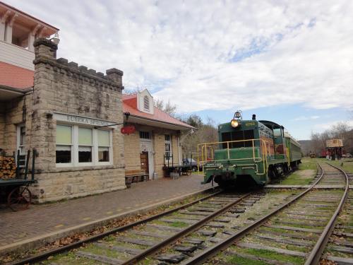 A green and yellow train at a historic station with stone architecture and tracks under a cloudy sky.