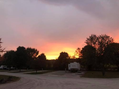 A serene sunset with vibrant orange and pink hues, framed by trees and a gravel road leading to an RV.