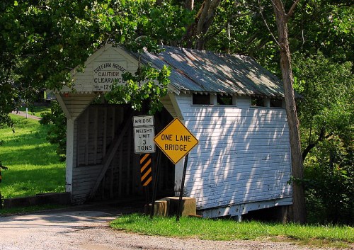 A weathered covered bridge with a caution sign and a one-lane bridge warning, surrounded by green trees.
