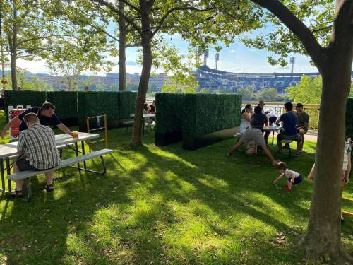 People enjoying a sunny day in a park with green hedges, tables, and a view of a stadium in the background.