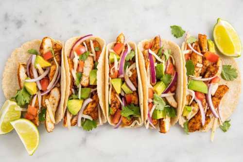 Five tacos filled with grilled chicken, tomatoes, onions, avocado, and cilantro, garnished with lime wedges.