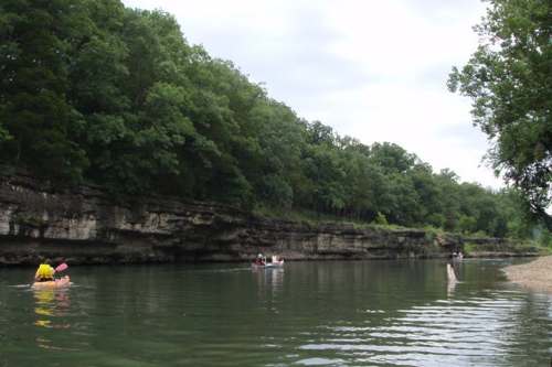 A serene river scene with people kayaking near rocky cliffs and lush green trees under a cloudy sky.