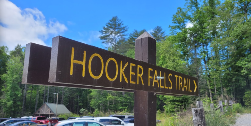 Sign for Hooker Falls Trail with a directional arrow, surrounded by trees and a parking area in the background.