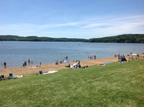A sunny beach scene with people swimming and relaxing on the sand by a calm lake, surrounded by green hills.