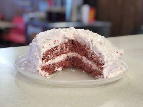 A slice of red velvet cake with cream frosting, partially cut and displayed on a clear plate.