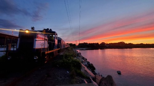 A train parked by a river at sunset, with vibrant colors reflecting in the water and a scenic sky.