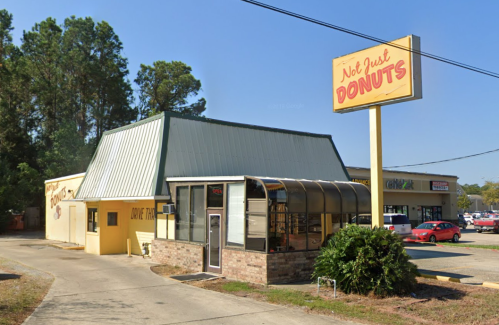 A yellow donut shop with a green roof and a large sign reading "Not Just Donuts," surrounded by trees and parking.