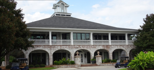 A large, two-story building with a gray roof and white columns, surrounded by greenery and golf carts parked in front.