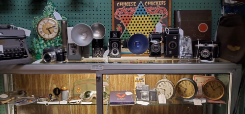 A display of vintage cameras, clocks, and a typewriter on a shelf, with a Chinese Checkers game in the background.