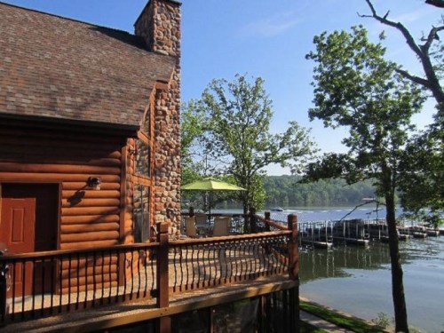 A log cabin with a stone chimney overlooks a serene lake, surrounded by trees and a dock in the background.