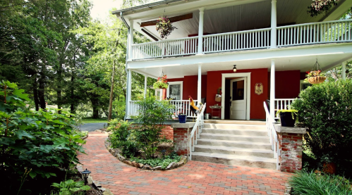 A charming two-story house with a porch, surrounded by greenery and a brick pathway leading to the entrance.