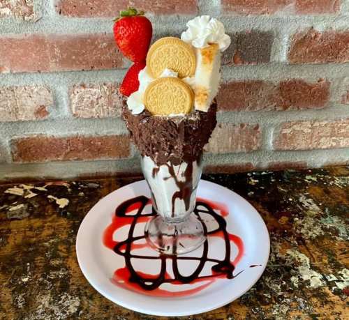 A tall glass filled with ice cream, topped with whipped cream, strawberries, cookies, and drizzled with chocolate and strawberry sauce.