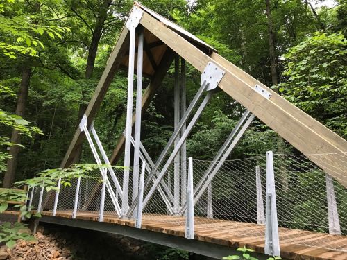 A modern wooden bridge with a triangular roof structure, surrounded by lush green trees in a forested area.