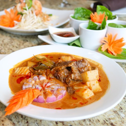 A bowl of rich, spicy curry with beef, potatoes, and onions, garnished with carrots, served with fresh herbs and sauces.