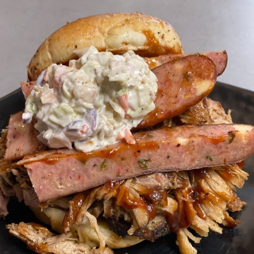 A hearty sandwich with pulled pork, sliced sausage, and coleslaw on a bun, drizzled with barbecue sauce.