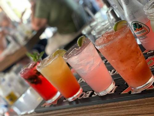 A row of colorful cocktails on a bar, featuring various fruits and garnishes, with blurred background elements.