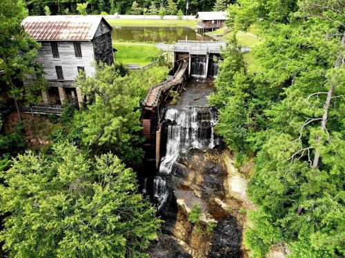 Aerial view of a rustic mill beside a waterfall, surrounded by lush greenery and a calm pond.