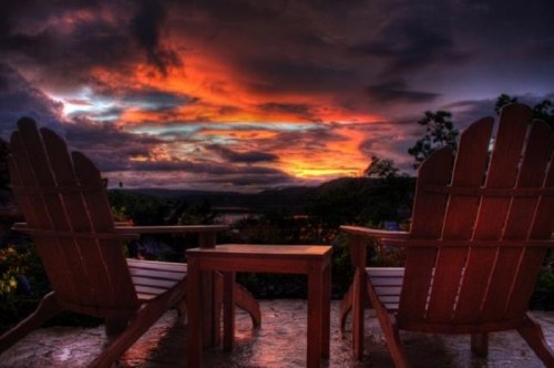 Two wooden chairs facing a vibrant sunset with dramatic clouds and a scenic landscape in the background.