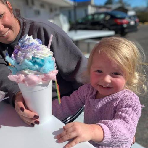 A smiling young girl holds a large, colorful ice cream cup topped with cotton candy, while an adult is beside her.