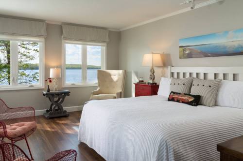 Bright bedroom with a large bed, armchair, and lake view through windows, featuring modern decor and warm lighting.