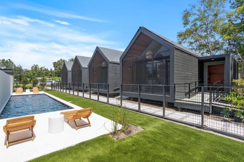 Modern cabins with large windows beside a swimming pool and landscaped lawn under a clear blue sky.