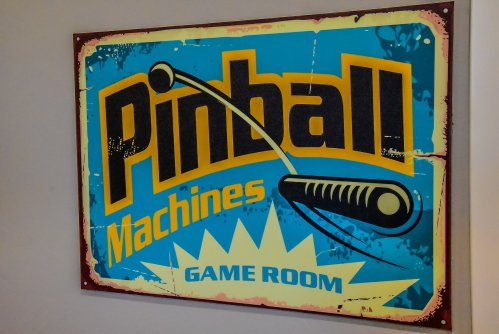 A vintage-style sign featuring "Pinball Machines" with a graphic of a pinball flipper, set in a game room theme.