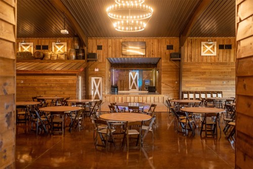 Interior of a rustic venue with wooden walls, round tables, and a stage area, featuring chandeliers and a warm ambiance.