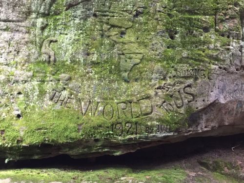 Carved inscription on a moss-covered rock, reading "THE WORLD U.S. 1921" with additional markings nearby.