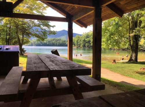 A serene lakeside view from a wooden pavilion, with a picnic table and people enjoying the water in the background.