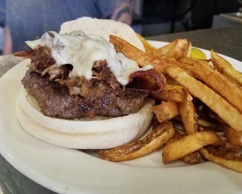 A juicy burger topped with pulled pork and mayo, served with crispy fries and a pickle on the side.