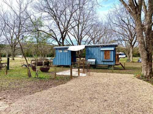 A rustic blue tiny house and shed in a grassy area, surrounded by bare trees and gravel pathways.