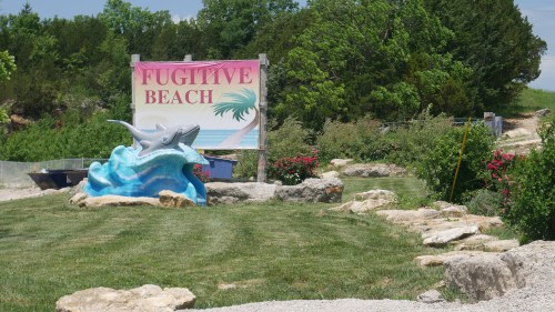 Sign for Fugitive Beach with a colorful dolphin sculpture, surrounded by greenery and flowers. Pathway leads into the area.