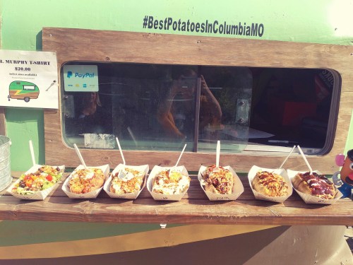 A variety of loaded potato dishes displayed in trays at a food stand, with a sign promoting the best potatoes in Columbia, MO.