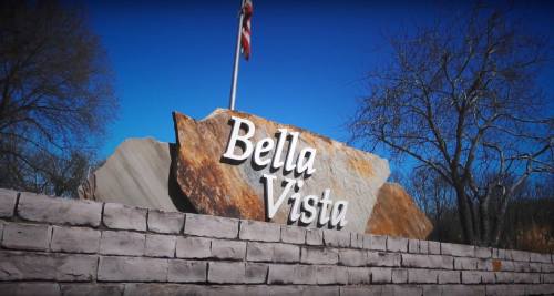 Sign reading "Bella Vista" on a stone structure with a clear blue sky in the background. An American flag is visible.