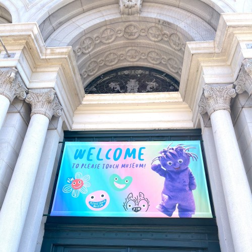 A colorful banner at a museum entrance reads "Welcome to Please Touch Museum!" with playful characters.