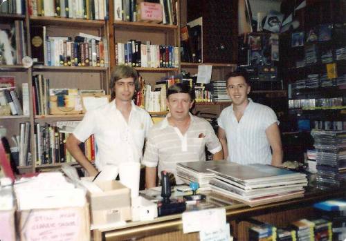 Three men stand behind a counter in a bookstore, surrounded by shelves of books and music CDs.