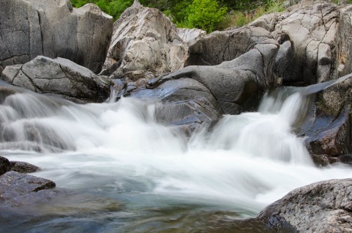 A serene river flows over smooth rocks, surrounded by lush greenery and rugged terrain.
