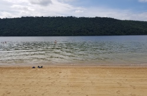 A sandy beach with calm water and a forested hillside in the background, under a partly cloudy sky.