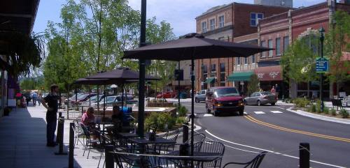 A sunny street scene with outdoor seating, trees, and shops, featuring cars and people enjoying the area.