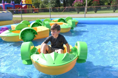 A young boy sits in a colorful paddle boat on a blue water surface, surrounded by other boats in a sunny outdoor setting.