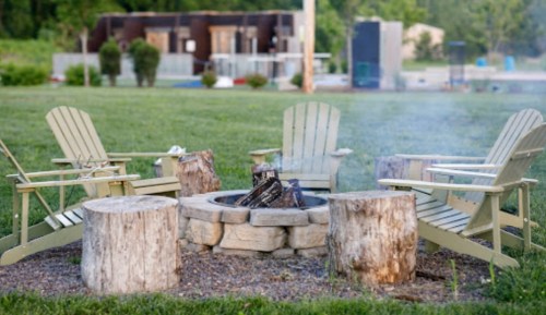 A cozy fire pit surrounded by wooden chairs and stumps, with smoke rising in a grassy area.