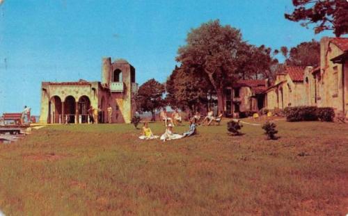 A sunny outdoor scene featuring people relaxing on a grassy area near rustic buildings and trees.