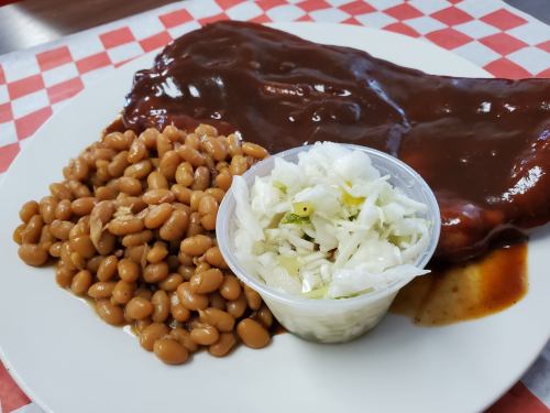 A plate featuring barbecue ribs with sauce, baked beans, and a side of coleslaw in a cup, on a checkered tablecloth.