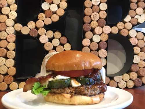 A close-up of a burger with bacon, lettuce, jalapeños, and sauce, served on a plate against a backdrop of cork letters.